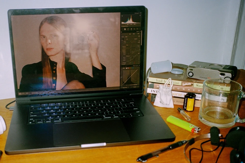 A laptop displays a portrait of a person in a black jacket taking a photo. The desk also features books, camera, film, and other objects.