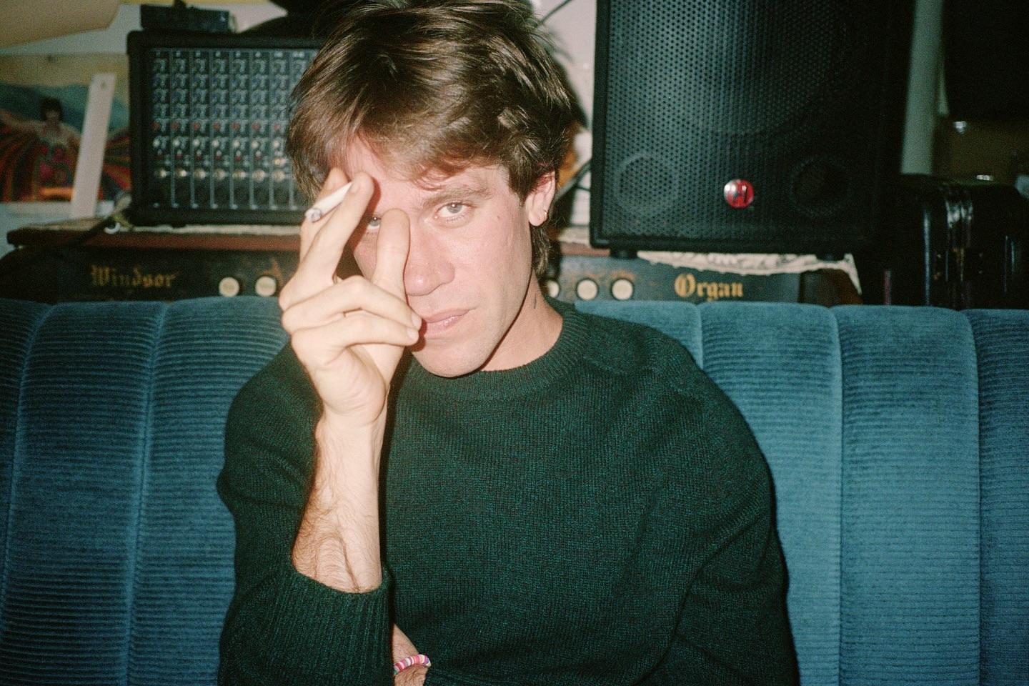 Portrait of an adult male with dark hair and a green sweater holding a cigarette, seated on a blue couch indoors.