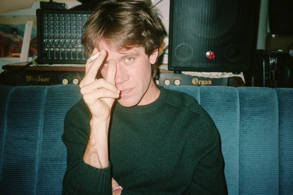Portrait of an adult male with dark hair and a green sweater holding a cigarette, seated on a blue couch indoors.