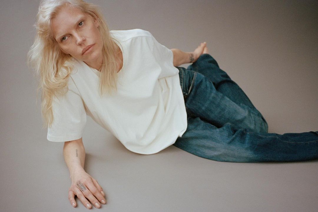 Woman with long blond hair poses while lying on a gray surface wearing a white t-shirt and blue jeans in a fashion portrait.