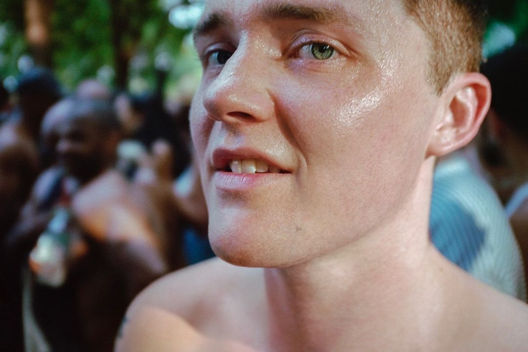 Close-up of a fair-skinned adult male with green eyes and light brown hair, with a crowd of people blurred in the background.