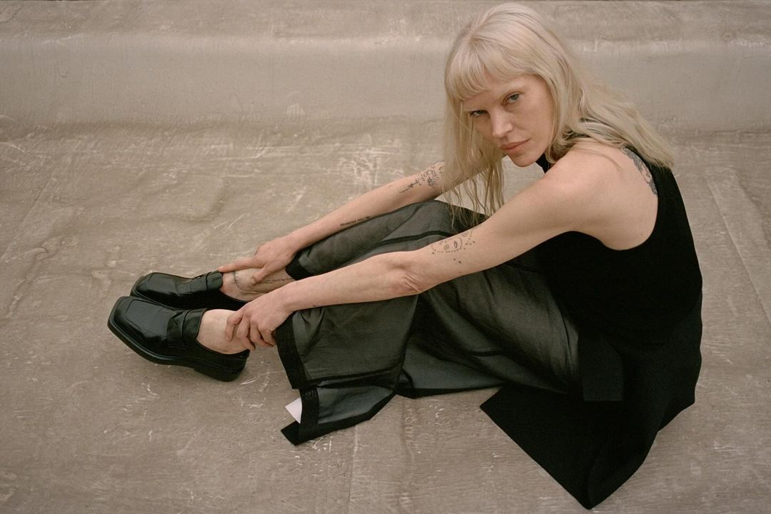 An adult female with fair skin and blonde hair is sitting on the floor wearing a black top, loose dark trousers, and shiny black shoes.
