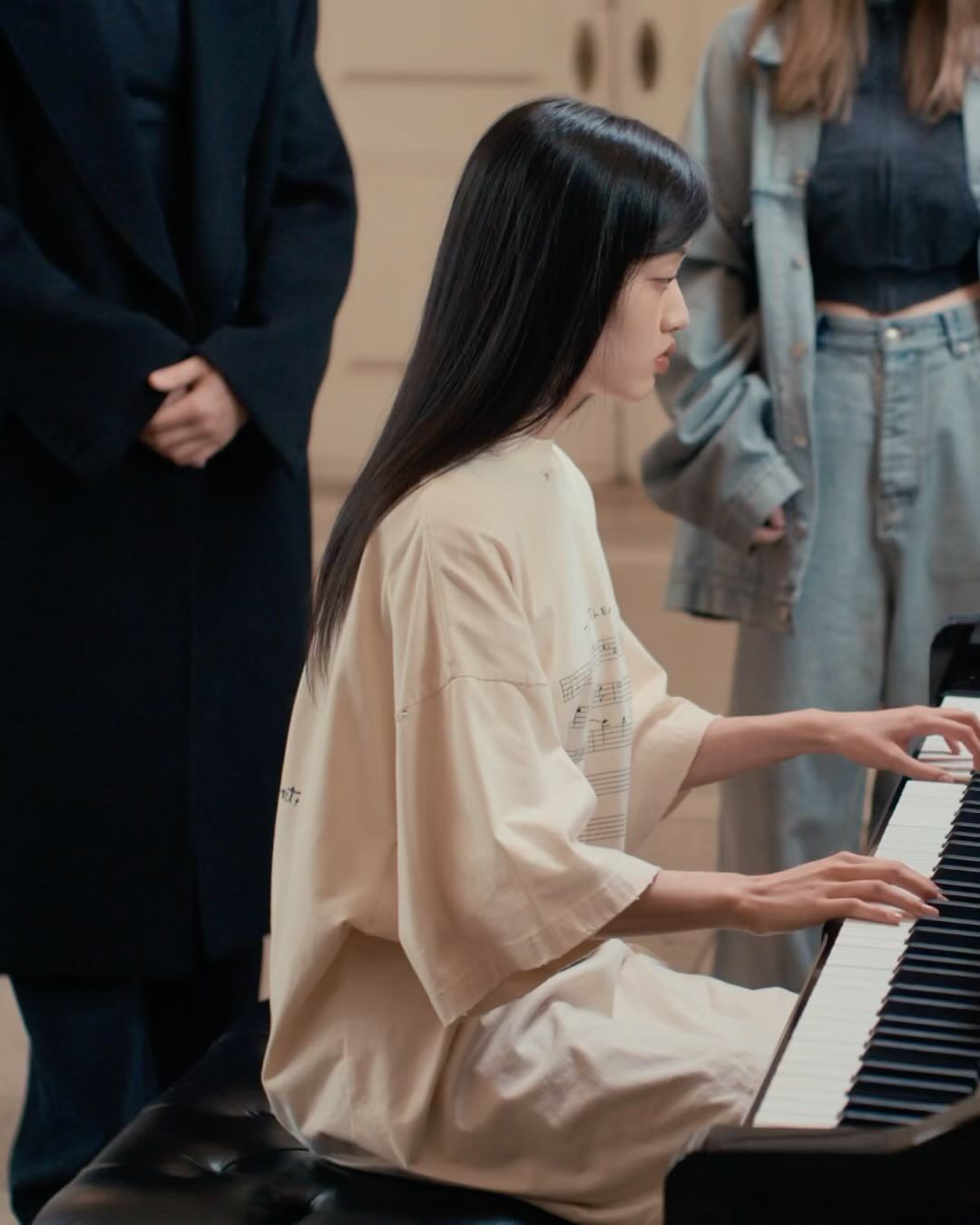 An Asian woman is playing a piano indoors while two other individuals stand in the background; she is wearing a beige top and sitting on a black bench.