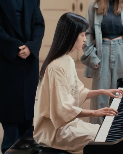 An Asian woman is playing a piano indoors while two other individuals stand in the background; she is wearing a beige top and sitting on a black bench.