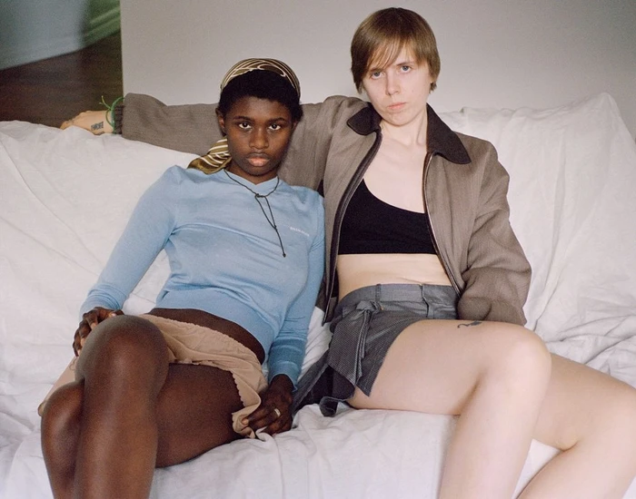Two young women seated together on a white bed, photographed by Sam Penn in New York.