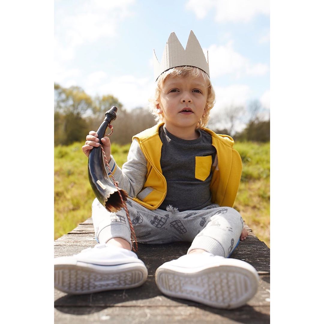 Young child with a cardboard crown sits outdoors holding a decorative horn, wearing a yellow vest and grey patterned pants.