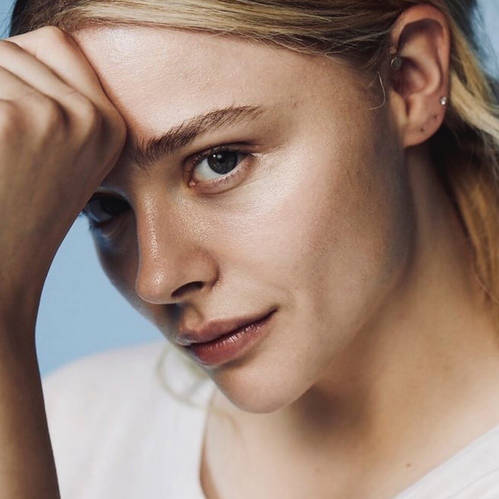Close-up portrait of a young woman with fair skin, blonde hair, and blue eyes looking at the camera with a neutral expression against a light blue background.