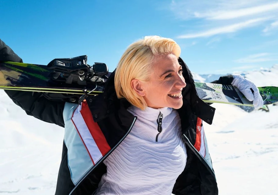 Smiling woman in ski wear carrying skis on shoulder against blue sky and snowy mountain backdrop, enjoying winter sports.