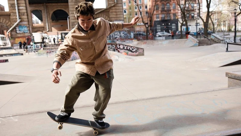 An Asian man riding skateboard in skate park, captured in medium shot, with urban buildings visible in background during daytime.