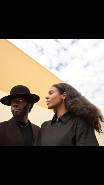 Portrait of an African American man in a hat standing with a woman wearing black against an abstract tan and blue sky backdrop.