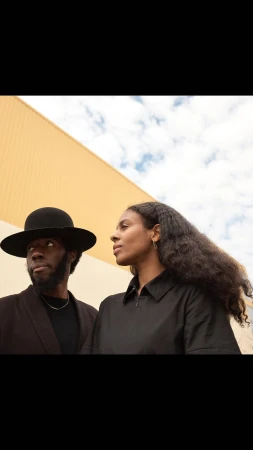 Portrait of an African American man in a hat standing with a woman wearing black against an abstract tan and blue sky backdrop.