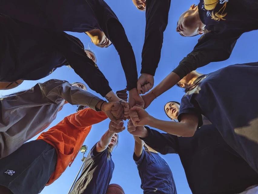 Low-angle shot of a diverse group of young adult athletes in sportswear with their fists together against a clear blue sky, teamwork.