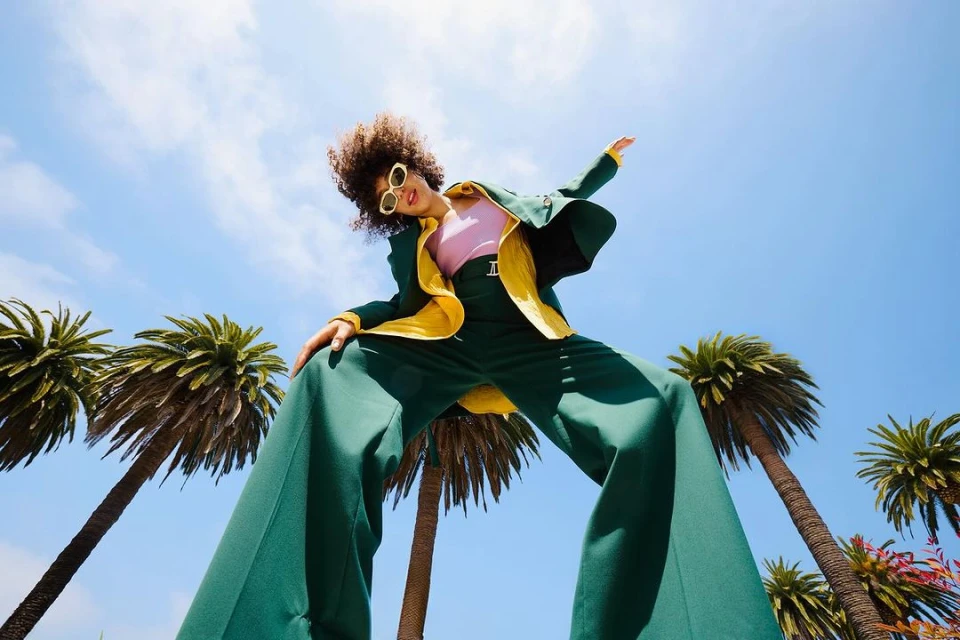 Woman in green suit poses under blue sky with palm trees. Retro style fashion shoot with wide-legged pants and vintage sunglasses. 
