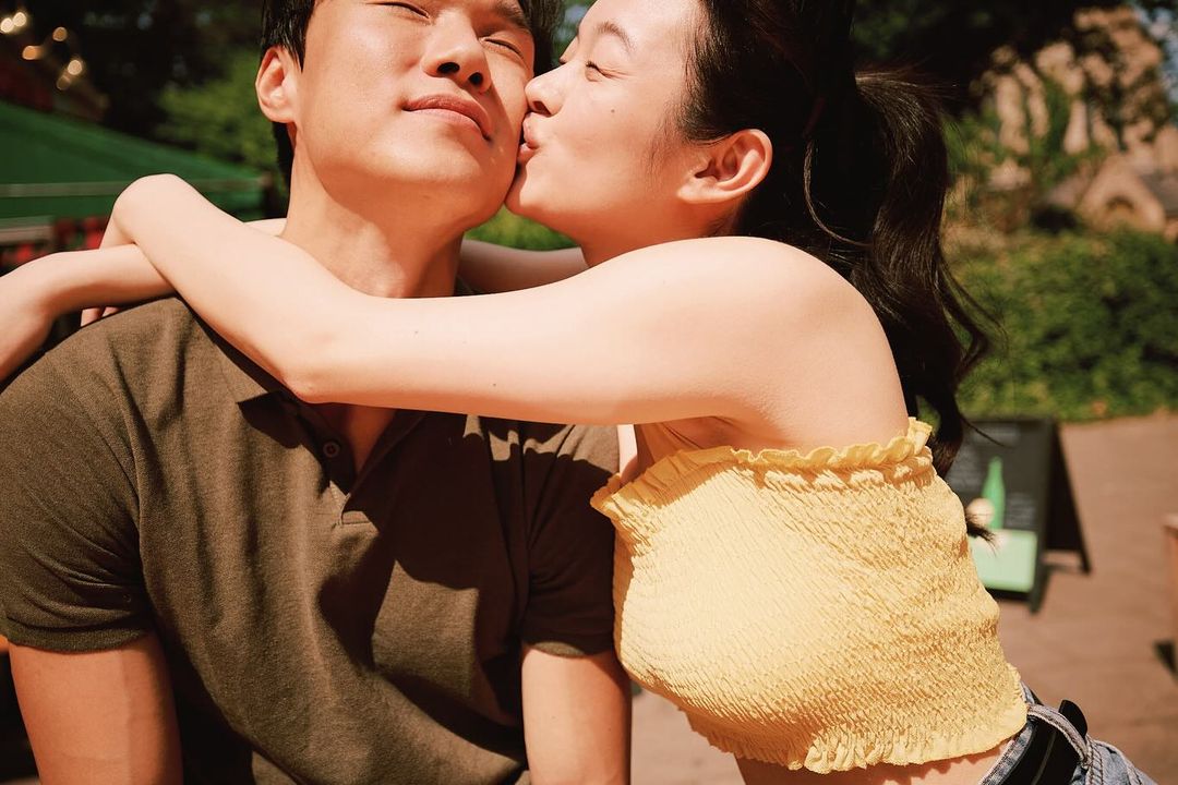 A smiling Asian couple embraces outdoors in sunlight. The woman kisses the man’s cheek while hugging him from behind near leafy foliage.