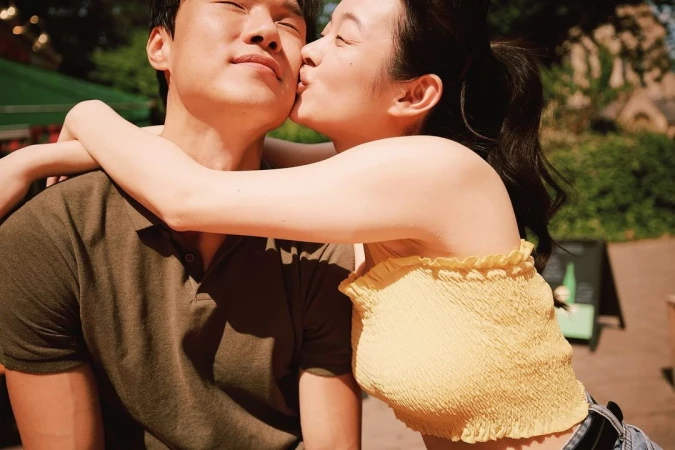 A smiling Asian couple embraces outdoors in sunlight. The woman kisses the man’s cheek while hugging him from behind near leafy foliage.