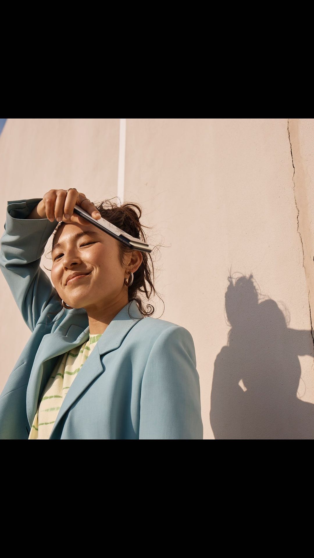 Smiling Asian woman in blue blazer balances tablet on her head in outdoor setting. Light brown wall serves as backdrop.