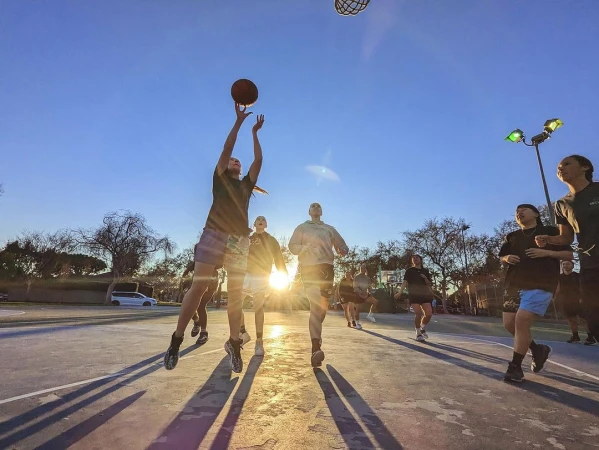 Adult female jumps to score basketball on outdoor court at sunset with team and sports lighting equipment in the background.