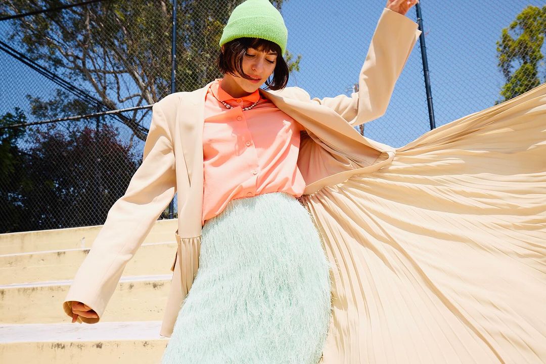 A stylish young woman poses on yellow stairs in a beige blazer, pastel pink shirt, mint green skirt, and a vibrant green beanie.