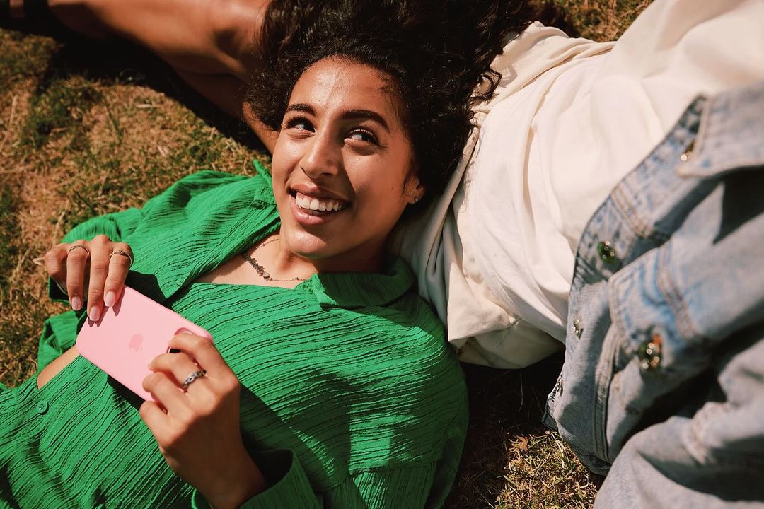 A smiling woman with curly hair in a green shirt lies on the grass, holding a pink mobile phone, alongside another person in a jacket.