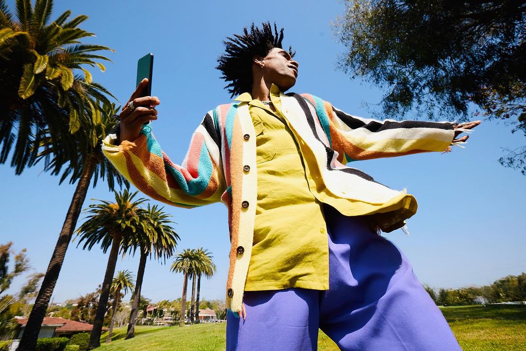 Adult male in a yellow shirt and colorful sweater stands in a sunny park with palm trees, holding a phone taking a selfie.