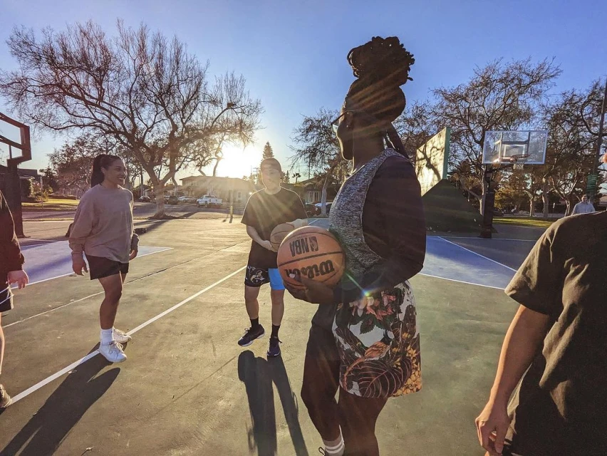 Group of young adults play basketball outdoors on a sunny day, dribbling and holding a Wilson NBA basketball on an outdoor court.