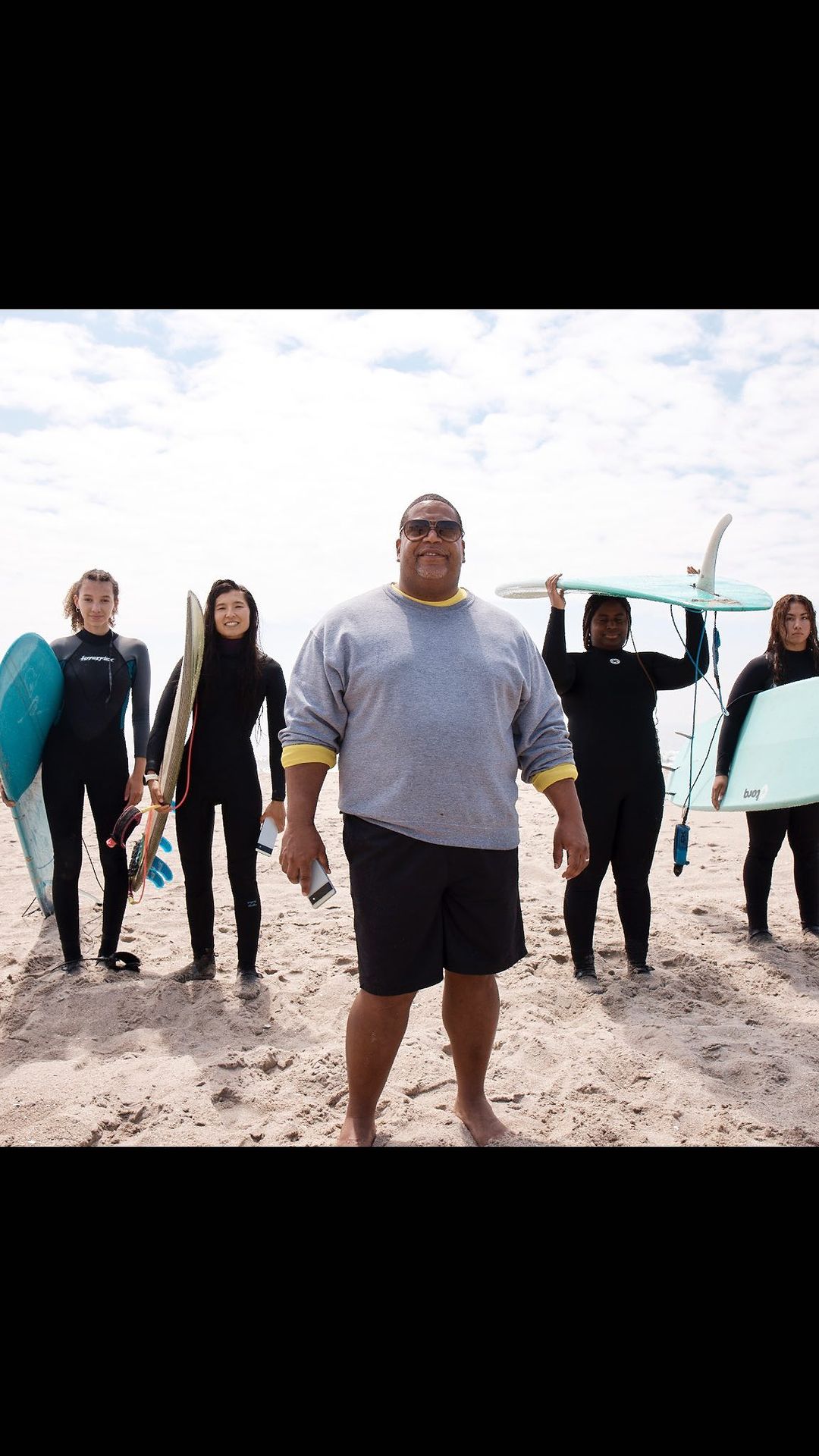 Group of five diverse adults with surfboards on a sandy beach, posing for the camera under a bright, sunny sky with few clouds.