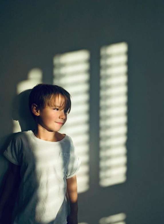 A young child in a white shirt stands indoors with sunlight casting window shade patterns on the wall and on his face.