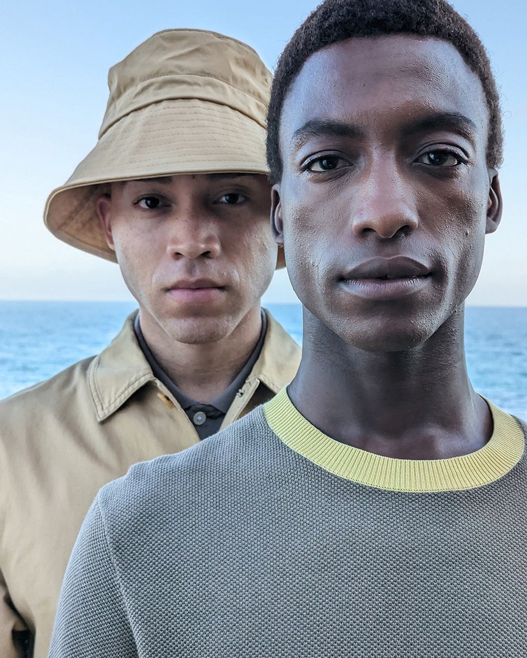 Two adult men pose in front of a bright blue ocean. One man is wearing a beige bucket hat, the other a grey sweater with a yellow neckline.