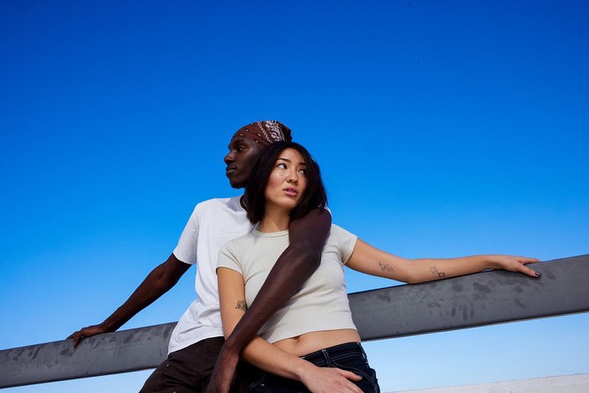 A young couple, one with a bandana, stands on a rooftop overlooking a clear blue sky. The man's arm is draped around the woman.
