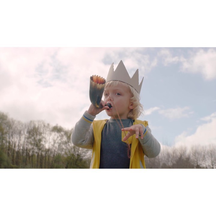 A young child with blond hair and a cardboard crown is blowing into a toy horn in a field with trees and a cloudy sky.