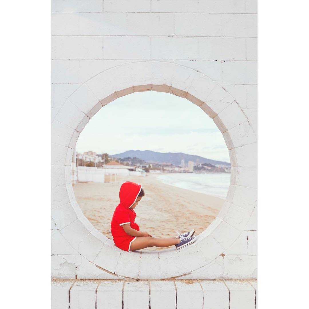 A child in a red hooded outfit sits in a round opening of a white wall, with a beach, ocean, and mountains visible in the background.