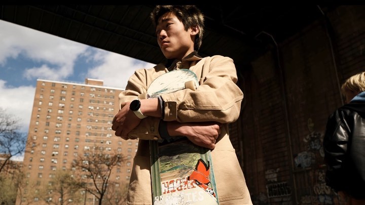 Low-angle view of an Asian man holding a skateboard in an urban area with a building and sky visible in the background, outdoor setting.