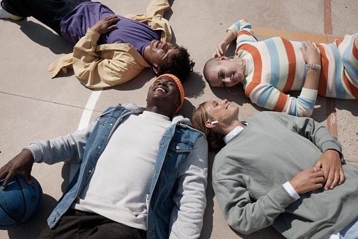 Overhead shot of four young adults laying in a circle on concrete, smiling with a basketball. Casual, diverse group enjoying time outdoors.