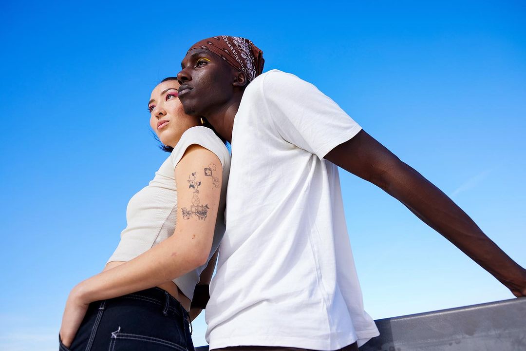 Two diverse adults in white t-shirts stand shoulder-to-shoulder under a vibrant blue sky, one with a bandana, looking off-frame.