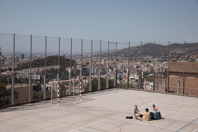 Four people sit on an outdoor concrete court with a soccer goal and a city skyline view on a clear day, enjoying a casual gathering.
