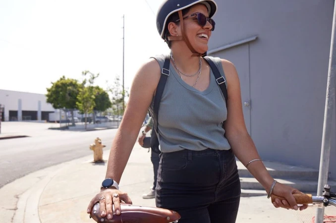 A smiling woman with a helmet rides a bicycle on an urban street, wearing sunglasses and a tank top on a sunny day near buildings.