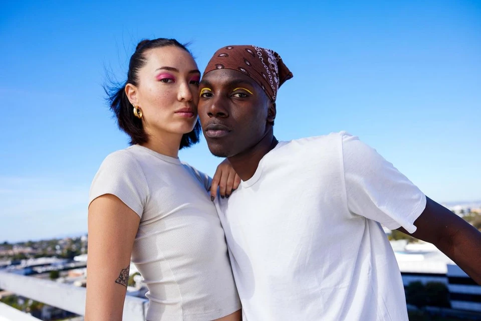 Portrait of two stylish people, a woman with pink eyeshadow and a man in a bandana with yellow eyeliner, posing outdoors against a blue sky.