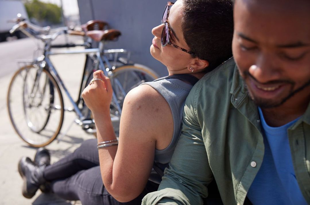 Two people, a woman in sunglasses and a man, sit back to back on a sunny city street with a bicycle parked behind them.