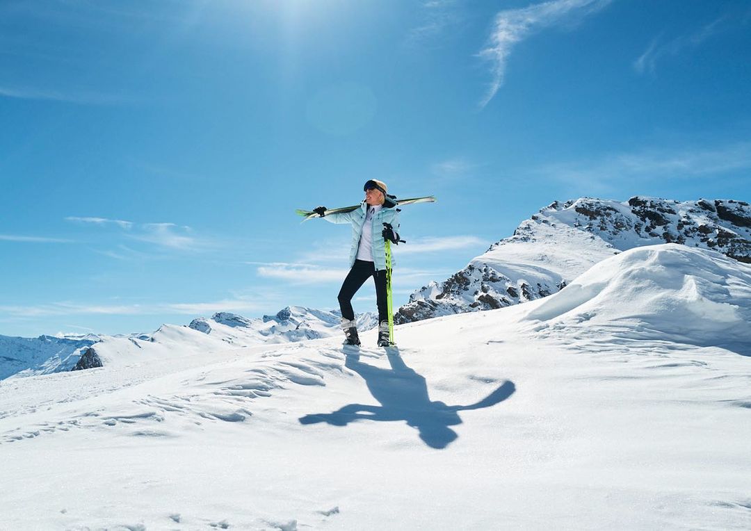 A woman on a snowy mountaintop holds skis in winter sportswear under bright blue sky with mountains in the distance on a sunny day.