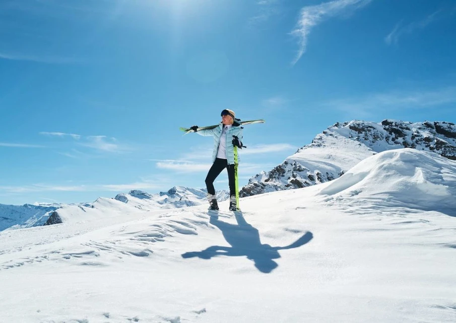 A woman on a snowy mountaintop holds skis in winter sportswear under bright blue sky with mountains in the distance on a sunny day.