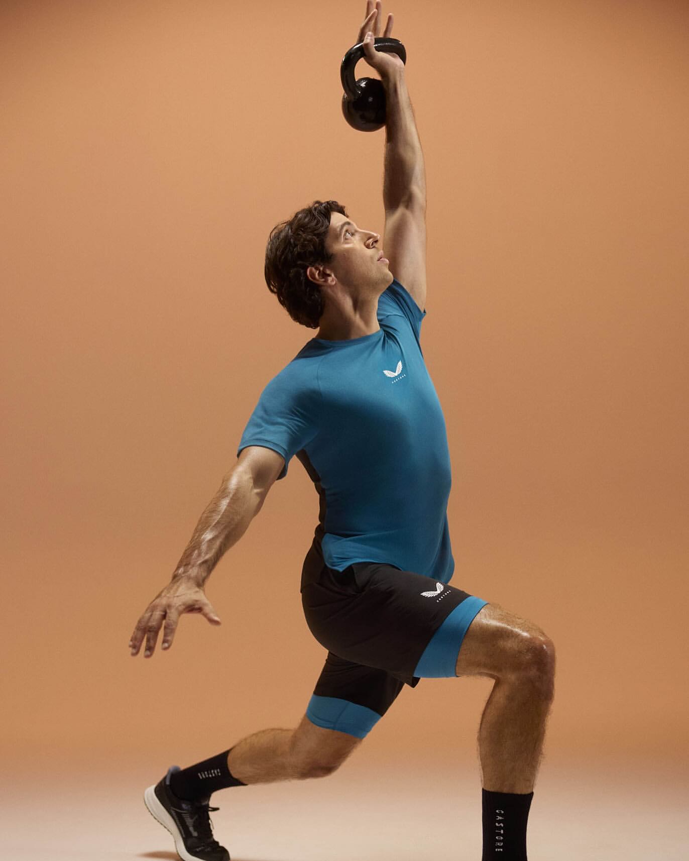 Athletic man in sportswear lunging with a kettlebell overhead against a neutral backdrop, promoting fitness and strength training.