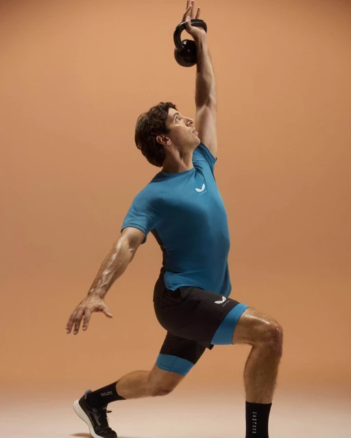 Athletic man in sportswear lunging with a kettlebell overhead against a neutral backdrop, promoting fitness and strength training.