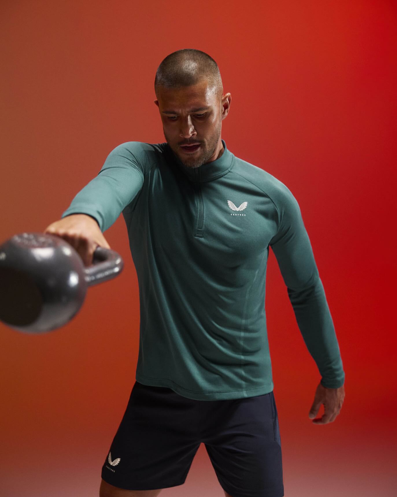 Adult male in athletic wear lifts a kettlebell in a studio with a red and orange gradient background.