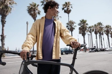 Young man with curly hair wearing a yellow overshirt and purple t-shirt standing outdoors next to a bicycle.
