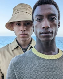 Two men standing outdoors by the ocean with a grey crewneck sweater and a tan bucket hat