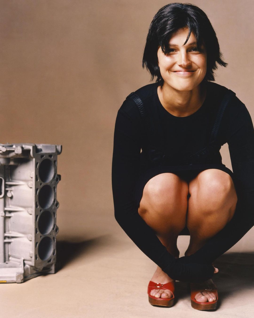 A woman squats next to a car engine block in a studio setting. She's wearing a black dress and red sandals, smiling at the camera.