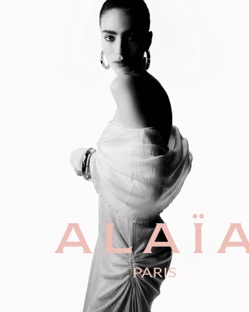 Fashion shot of a woman with hoop earrings wearing a white dress and shawl with Alaïa Paris text on a white background.