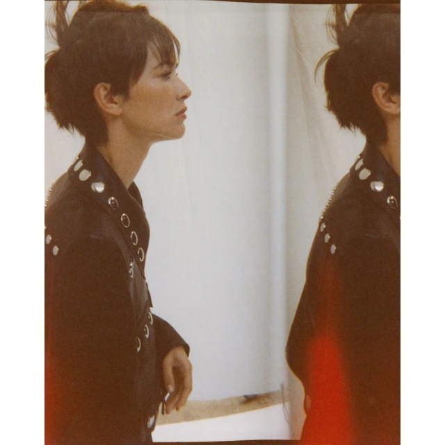 A brunette woman in a black leather jacket poses in profile view, duplicated in a shallow depth-of-field fashion photograph indoors.