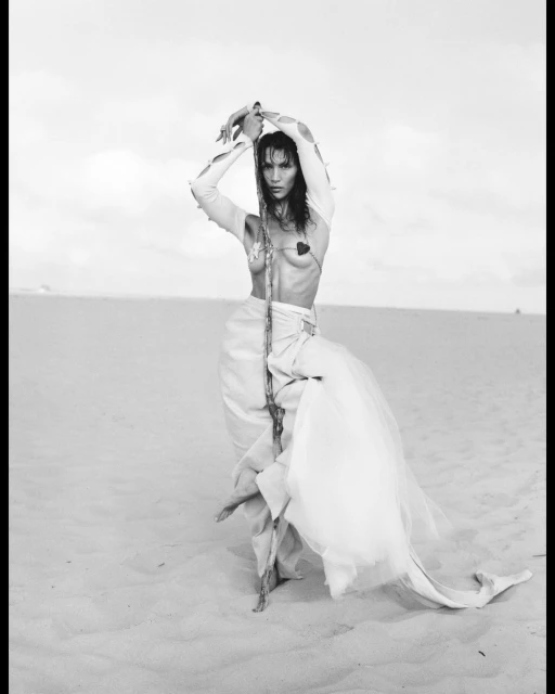 Monochrome image featuring a woman with bare chest in a desert setting, holding a staff and adorned with white skirt-like fabric.