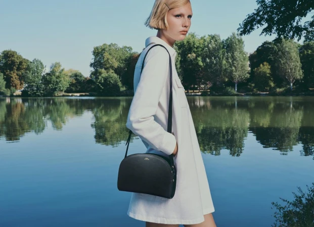 Model in white shirt dress carrying a black crossbody handbag standing near a reflective lake with trees